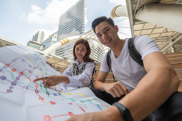 Jóvenes y lindos viajeros asiáticos en pareja dan un paseo juntos para hacer turismo en la gran ciudad y en busca de papel mapa encontrar destinos y atracciones turísticas con edificios altos en el fondo. - Foto, Imagen