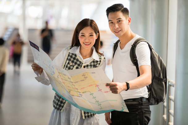 Couple travelers holding a paper map in hands together and looking and finding destination and station for lines of a sky train with a blurry background. Idea for tourists travels on vacation. - Photo, Image