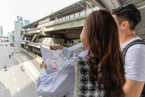 Couple travelers holding a paper map in hands together and looking and finding destination and station for lines of a sky train in a big city. Idea for tourists travels on vacation. - Photo, Image