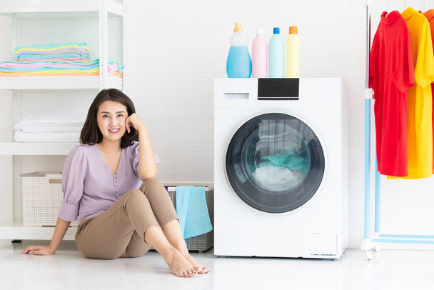 Lächelnde asiatische Frau, die auf dem Boden sitzt und mit einem Gefühl der Entspannung in eine Kamera blickt, während sie auf eine Waschmaschine wartet, die sie putzt - Foto, Bild