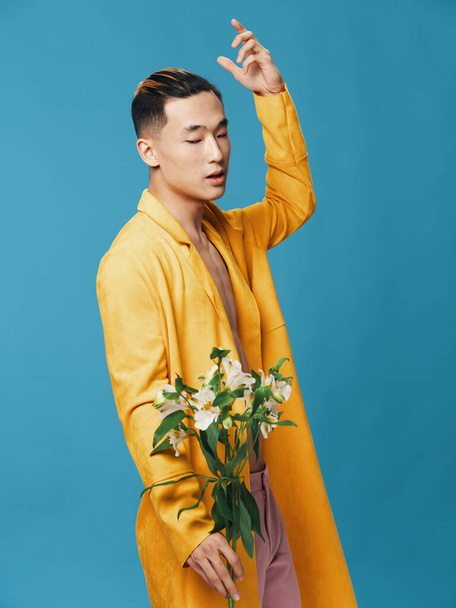 Молодой человек азиатского вида в желтом пальто с букетом цветов жестикулирует руками - Фото, изображение