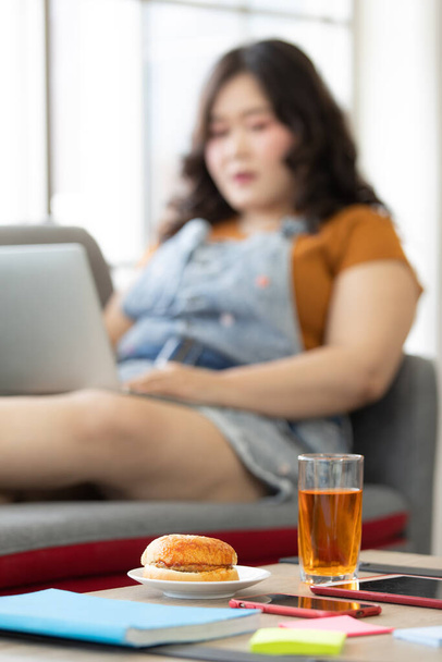 白い皿にハンバーガーの写真と、食べ物を探して自宅のソファに座っている太った女性の背景がぼやけているテーブルの上のジュースのガラス。ハンバーガーの選択的フォーカス. - 写真・画像