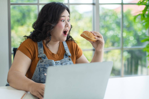 Portrét tlusté asijské ženy, která sedí a drží hamburger, otevírá ústa a váhá, zda má jíst, zatímco v místnosti používá notebook. Zdravý a práce z domova koncept. - Fotografie, Obrázek