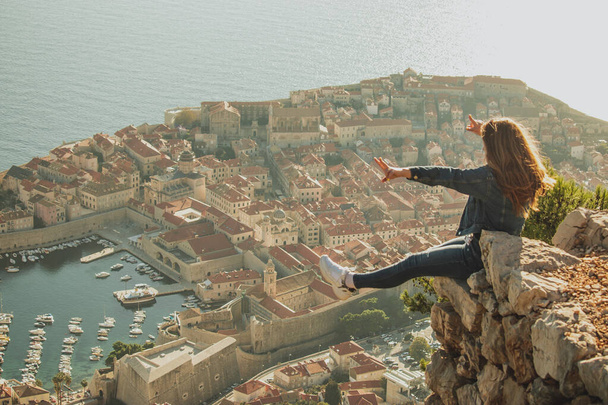Feliz chica emocionada sentada en el borde de una roca con la vieja ciudad de Dubrovnik detrás de ella. Hermosa ciudad antigua que brilla durante una puesta de sol por la tarde, visitando durante la temporada covid - Foto, Imagen
