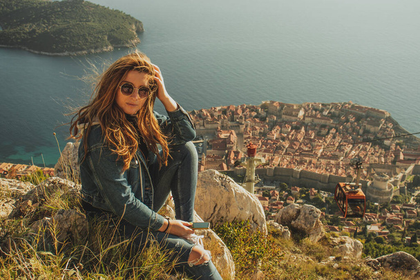 Attractive brunette sitting on the Srd hill above the city of Dubrovnik, observing the city in the distance. Wind blowing her hair above the ancient famous landmark town - Фото, изображение