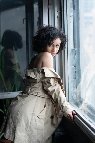 young pretty woman stands near the window in a raincoat - Foto, Imagen