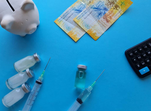 dinheiro suíço, mealheiro, calculadora, frasco de remédio e seringa no fundo azul - Foto, Imagem