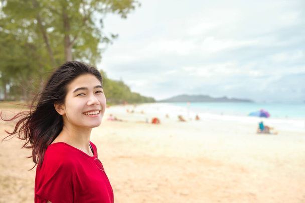 Smiling biracial Asian Caucasian teen girl at tropical beach looking out towards aqua blue ocean on cloudy day - Foto, afbeelding