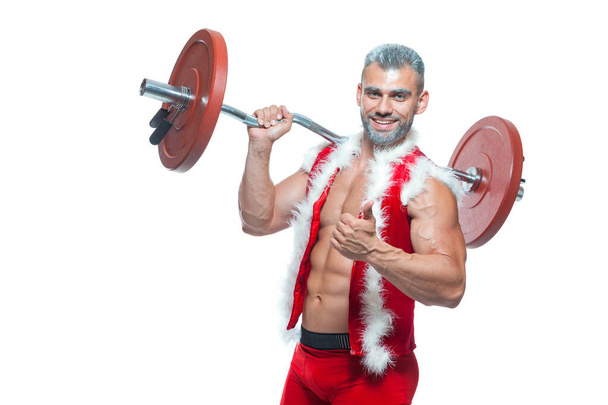 Sexy Santa Claus. Bodybuilder young handsome santa clause smile holds a barbell with red pancakes on the shoulder and shows off abs cubes at New Years eve and Christmas winter holiday white background - Foto, afbeelding
