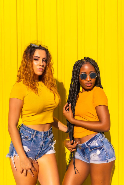 Girlfriends lifestyle, black girl with long braids and blonde Caucasian in yellow shirts and short jeans on a yellow background. Urban session with warm colors in the city - Fotografie, Obrázek