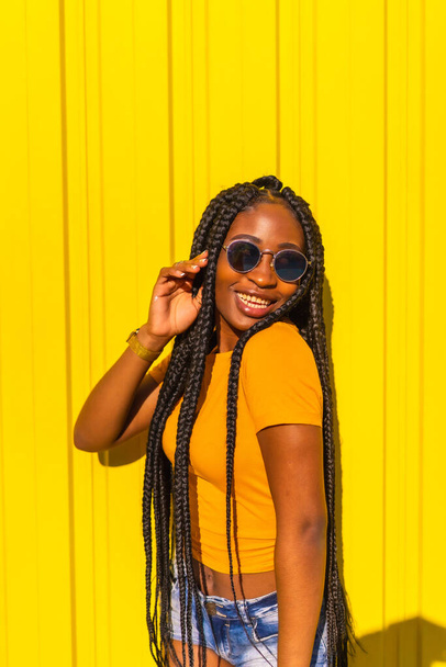 Lifestyle, attractive black girl with long braids, yellow t-shirts and short jeans on a yellow background. With sunglasses having fun, trap dancer - Fotoğraf, Görsel