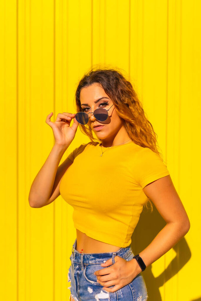 Lifestyle, Caucasian blonde girl in yellow shirts on a yellow background. Fashionable posing putting on the gorgeous sunglasses with a smile - Фото, изображение
