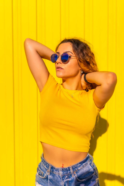 Lifestyle, Caucasian blonde girl in yellow shirts on a yellow background. Fashionable pretty girl posing in a calm expression, wearing sunglasses - Фото, изображение