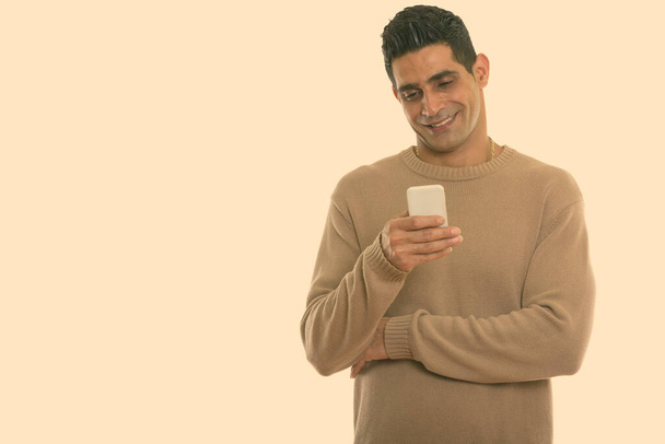 Studio shot van jonge gelukkig Perzische man glimlachen tijdens het gebruik van mobiele telefoon - Foto, afbeelding