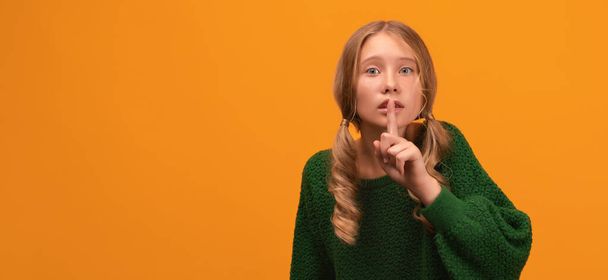 Portret van een mooi blond meisje van 12-14 jaar in een warmgroene trui met wijsvinger op de lippen. Stilte en geheim concept. Studio schot, gele achtergrond, geïsoleerd. Gezichtsuitdrukking - Foto, afbeelding