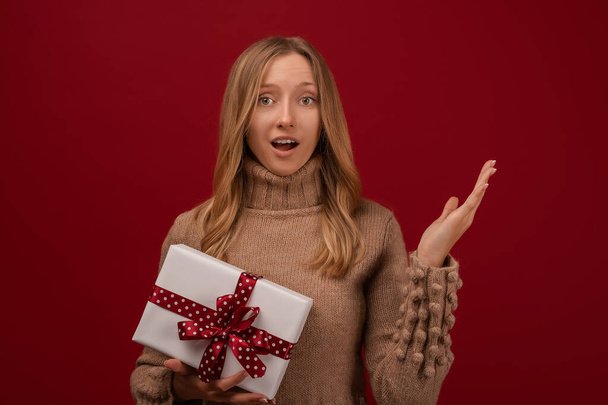 Изображение очаровательной молодой блондинки в модном вязаном свитере, улыбающейся и держащей в руках подарок с красной лентой. Студия сняла красный фон. Концепция новогодних праздников - Фото, изображение