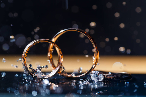 Goldene Trauringe für Brautpaare an einem Hochzeitstag auf schwarzem Hintergrund mit Wassertropfen. Schmuck - Foto, Bild