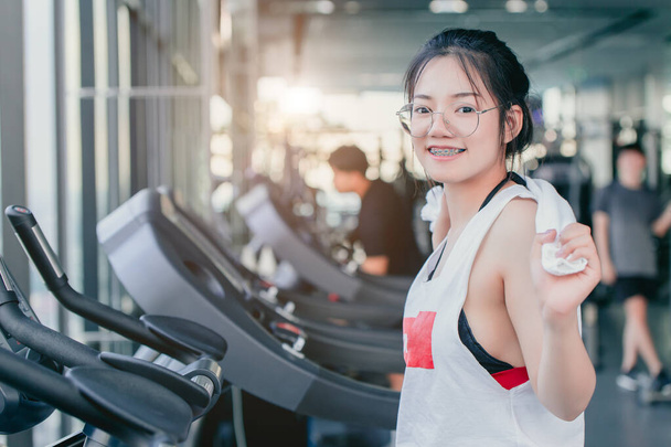 Aziatisch tiener beugel meisje cardiotraining oefening in sport club met handdoek op haar schouders glimlachen na het sporten op loopband in de sportschool op zoek camera. - Foto, afbeelding