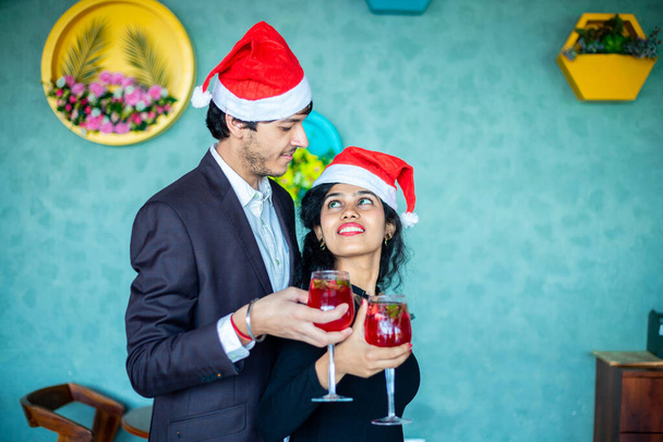 Boldog fiatal pár télapó kalapot visel, együtt ünneplik a karácsonyt, miközben piros koktélpoharakat tartanak, nézik egymást otthon, másolják a helyet. Ünnepi koncepció. - Fotó, kép