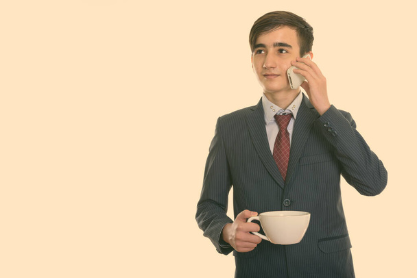 Studio shot of young handsome Persian teenage businessman thinking while holding coffee cup and talking on mobile phone - Photo, Image