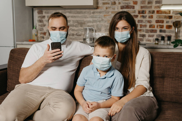 Father, mother and son are sitting on a sofa in medical face masks to avoid the spread of coronavirus (COVID-19). Family during quarantine at home. Dad is reading news on a phone near relatives - Photo, Image