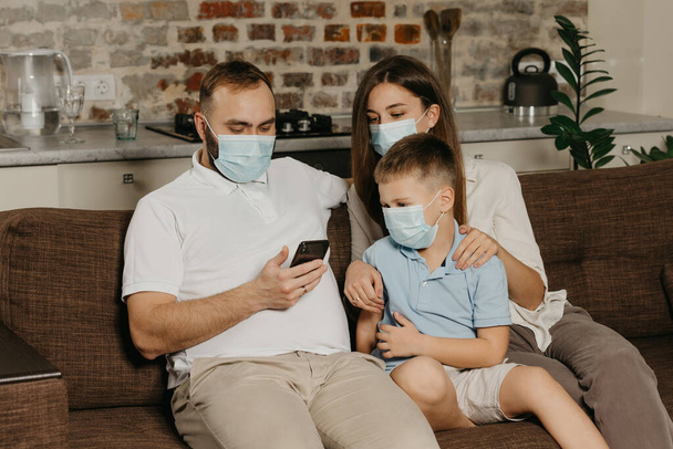 Батько, мати і син сидять на дивані в масках для медичних осіб, щоб уникнути поширення коронавірусу (COVID-19). Родина під час карантину вдома. Тато дивиться новини по телефону біля своїх родичів. - Фото, зображення