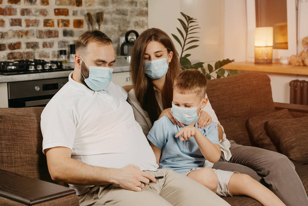 Отец, мать и сын сидят на диване в медицинских масках, чтобы избежать распространения коронавируса (COVID-19). Семья во время карантина дома. Родственники смотрят видео по мобильному телефону на диване - Фото, изображение