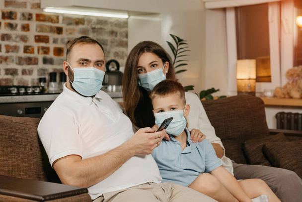 Father, mother and son are sitting on a sofa in medical face masks to avoid the spread of coronavirus (COVID-19). Family during quarantine at home. Relatives are posing on the couch - Photo, Image