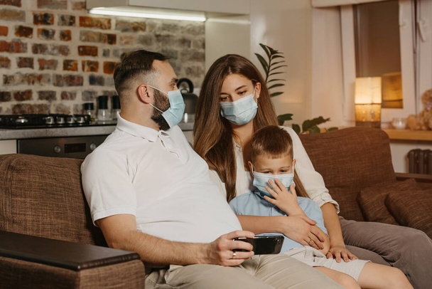 Πατέρας, μητέρα και γιος κάθονται σε έναν καναπέ με ιατρικές μάσκες προσώπου για να αποφευχθεί η εξάπλωση του coronavirus (COVID-19). Οικογένεια σε καραντίνα στο σπίτι. Ο μπαμπάς δείχνει βίντεο στους συγγενείς του. - Φωτογραφία, εικόνα