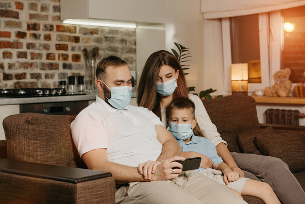 Père, mère et fils sont assis sur un canapé masqué pour éviter la propagation du coronavirus (COVID-19). Famille pendant la quarantaine à la maison. Les parents apprécient le contenu sur le téléphone portable dans la soirée - Photo, image