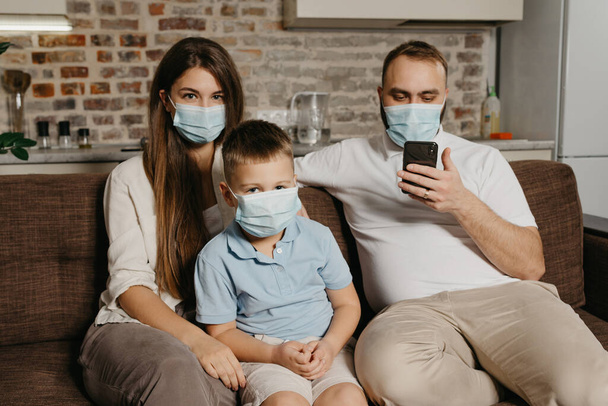 Father, mother and son are sitting on a sofa in medical face masks to avoid the spread of coronavirus (COVID-19). Family during quarantine at home. Dad is reading news on a phone near relatives - Fotoğraf, Görsel