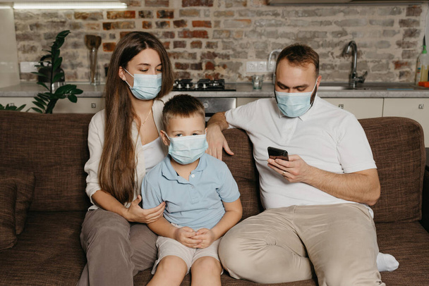Батько, мати та їх син сидять на дивані в медичних масках для обличчя, щоб уникнути поширення коронавірусу (COVID-19). Сім'я під час карантину вдома. Тато читає новини по телефону поблизу родичів
 - Фото, зображення