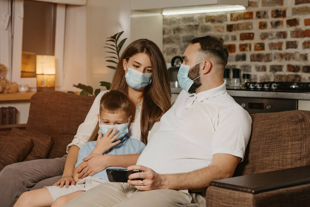 Ojciec, matka i syn siedzą na kanapie w maskach medycznych, aby uniknąć rozprzestrzeniania się koronawirusu (COVID-19). Rodzina podczas kwarantanny w domu. Tata demonstruje film swoim krewnym. - Zdjęcie, obraz