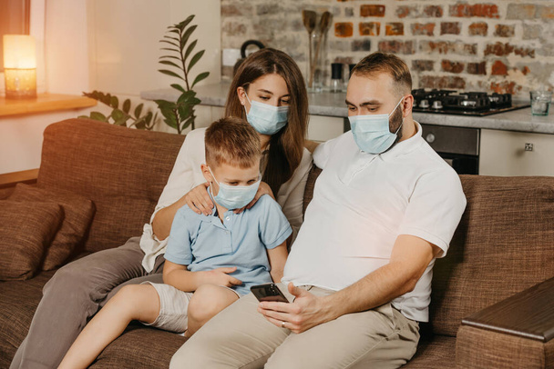 Ojciec, matka i syn siedzą na kanapie w maskach medycznych, aby uniknąć rozprzestrzeniania się koronawirusu (COVID-19). Rodzina podczas kwarantanny w domu. Krewni oglądają wiadomości przez komórkę na kanapie. - Zdjęcie, obraz