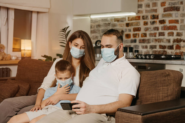 Πατέρας, μητέρα και γιος κάθονται σε έναν καναπέ με ιατρικές μάσκες προσώπου για να αποφευχθεί η εξάπλωση του coronavirus (COVID-19). Οικογένεια σε καραντίνα στο σπίτι. Συγγενείς απολαμβάνουν το περιεχόμενο σε ένα smartphone - Φωτογραφία, εικόνα