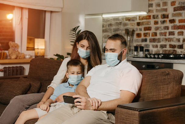 Otec, matka a syn sedí na pohovce v obličejových maskách, aby se zabránilo šíření koronaviru (COVID-19). Rodina během karantény doma. Příbuzní si večer užívají obsah mobilního telefonu - Fotografie, Obrázek