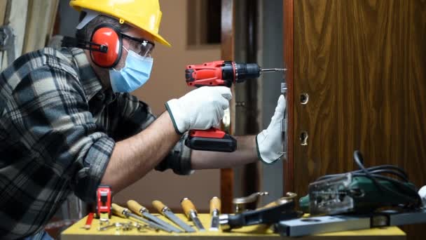 作業修理で大工の労働者と部屋のドアロックをインストールし、コロナウイルス感染を防ぐために外科用マスクを着用してください。職場でのパンデミックコビト-19の防止。大工さん。映像. - 映像、動画