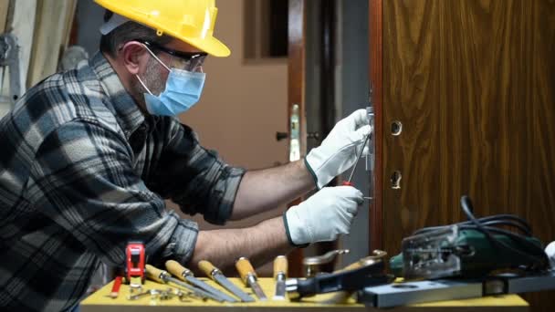 Carpenter worker at work repair and install a room door lock, wear the surgical mask to prevent Coronavirus infection. Preventing Pandemic Covid-19 at the workplace. Carpentry. Footage. - Footage, Video