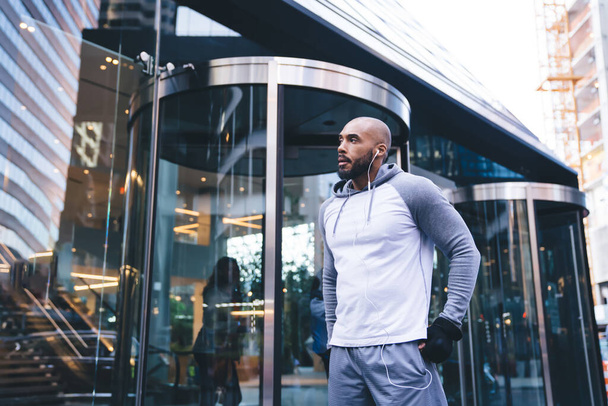 Bald African American sportsman standing at street near glass entrance to building during break in training and listening music using headphones - Fotó, kép