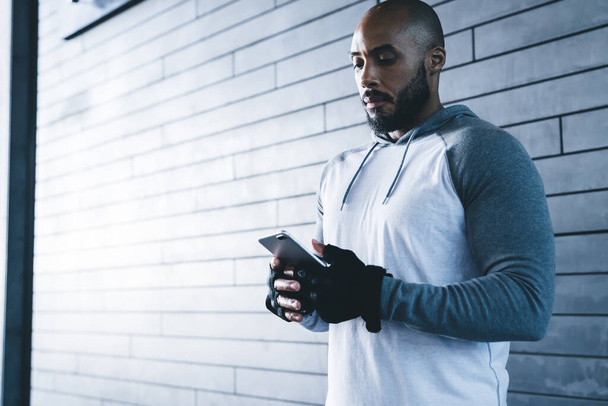 Bearded sporty black male in gloves browsing smartphone while standing alone against gray brick wall in modern fitness gym in daylight - Фото, изображение