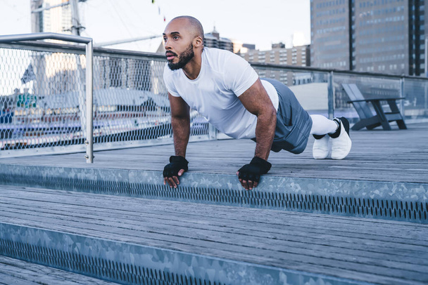 Side view of active adult black bodybuilder in tracksuit and gloves having morning workout with high plank pose on bridge looking away on background of buildings - Fotoğraf, Görsel