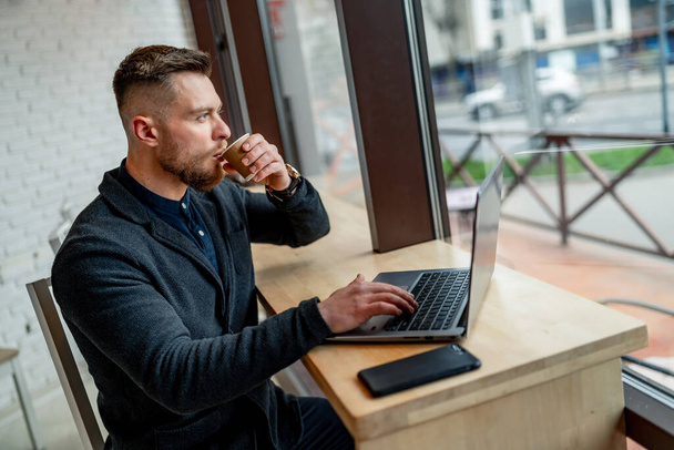 Joven hombre de negocios bebiendo café y trabajando en un portátil en un café. hombre guapo en traje oscuro. - Foto, Imagen