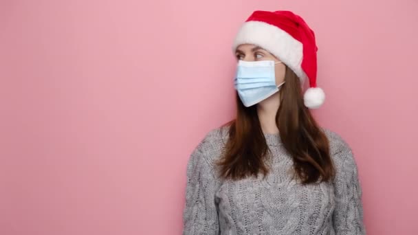Jonge vrouw met medisch masker punten met de hand opzij, toont kopieerruimte voor reclame, draagt Santa Kerstmis hoed en winter trui, geïsoleerd op roze studio muur. Covid-19 en Nieuwjaarsconcept - Video
