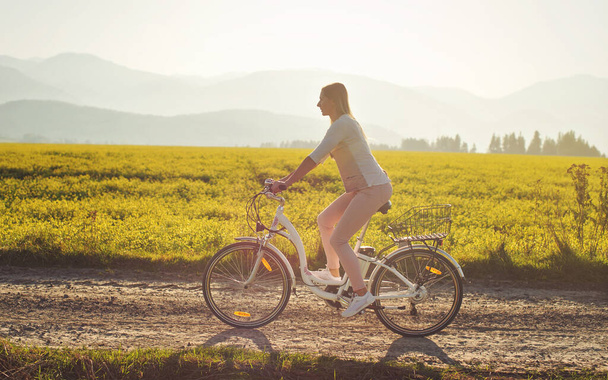 Young woman rides electric bike over dusty country road, strong afternoon sun backlight in background shines on yellow flowers field, view from side - Foto, Imagem