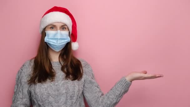 Young woman in medical face mask keeps palm raised, points with forefinger against pink wall for advertisement, dressed in Christmas hat and winter sweater. Pandemic coronavirus and new year concept - Imágenes, Vídeo