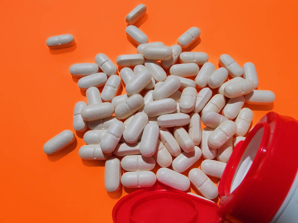 Witte versnipperde pillen en witte pot met rode deksel op oranje achtergrond. Selectieve focus. Het begrip voedingssupplementen. - Foto, afbeelding