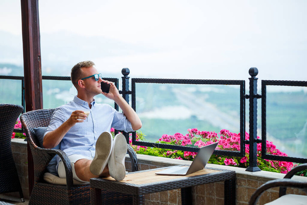 Geschäftsmann, der im Urlaub an einem Laptop mit schönem Panoramablick arbeitet. Erfolgreicher Manager trinkt Kaffee und telefoniert auf Reisen mit Kaffee - Foto, Bild