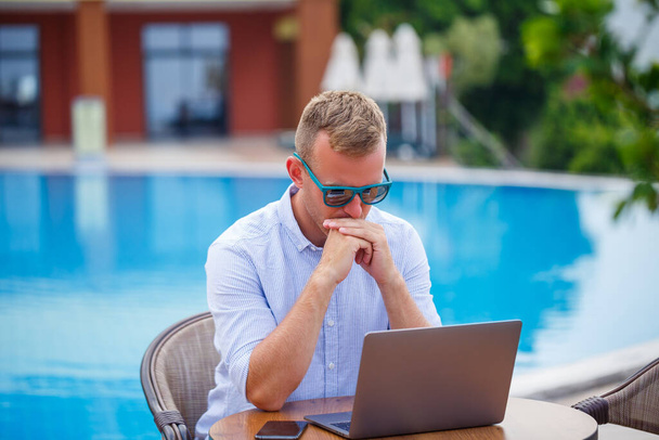 Jovem homem de negócios em óculos de sol trabalha em um laptop sentado em uma mesa perto da piscina. Trabalho remoto. Freelancer - Foto, Imagem