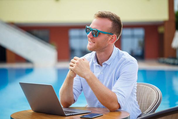 Επιτυχημένος όμορφος άντρας επιχειρηματίας με γυαλιά ηλίου δουλεύει σε ένα λάπτοπ δίπλα στην πισίνα. Απομακρυσμένη δουλειά. Ελεύθερος επαγγελματίας - Φωτογραφία, εικόνα