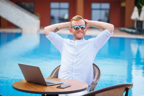 Un bel homme d'affaires avec des lunettes de soleil travaille sur un ordinateur portable près de la piscine. Travail à distance. Indépendante - Photo, image